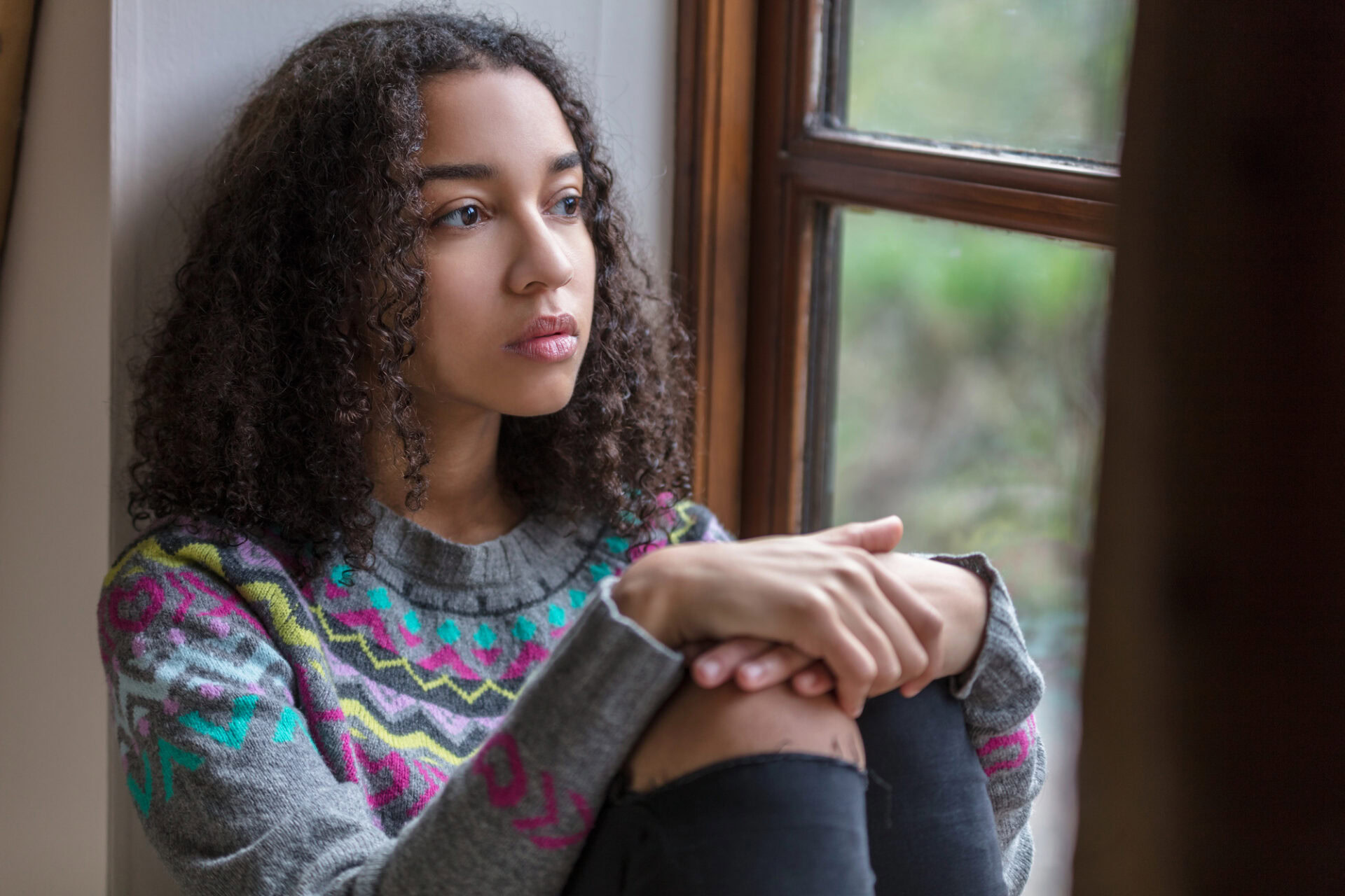 Teen girl looking out window, feeling worried about substance abuse and mental health.