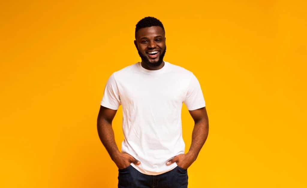Bright smiling man with hands in pockets, casual white t-shirt, vibrant yellow background for positive vibe.