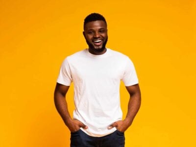 Bright smiling man with hands in pockets, casual white t-shirt, vibrant yellow background for positive vibe.