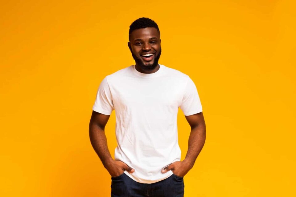 Bright smiling man with hands in pockets, casual white t-shirt, vibrant yellow background for positive vibe.