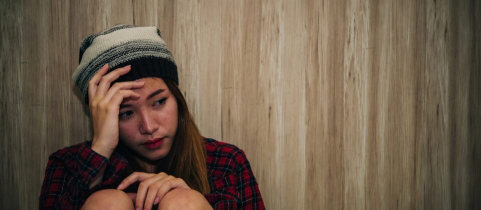 Anxious young woman in a beanie and plaid shirt sitting against a wooden wall.