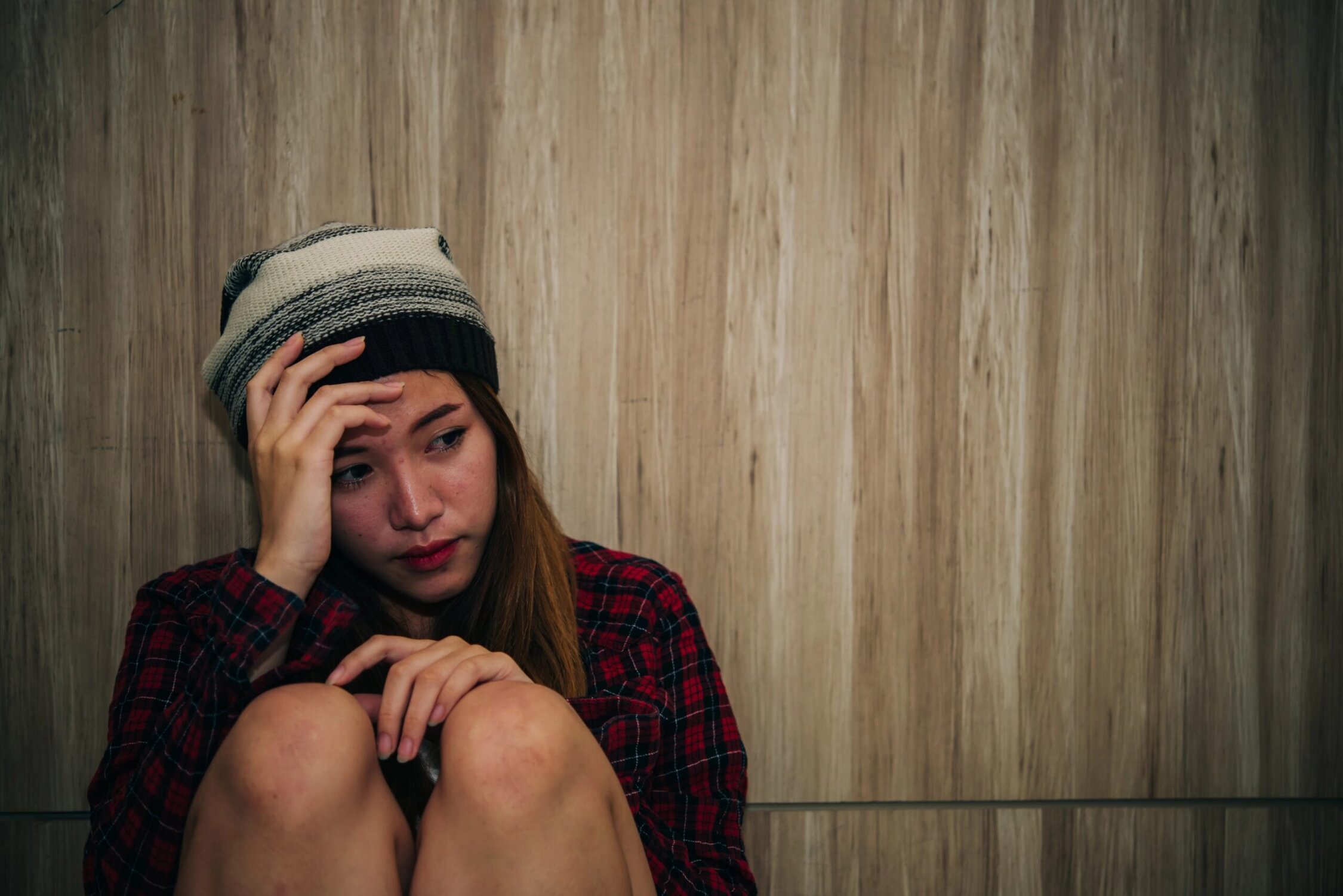 Anxious young woman in a beanie and plaid shirt sitting against a wooden wall.
