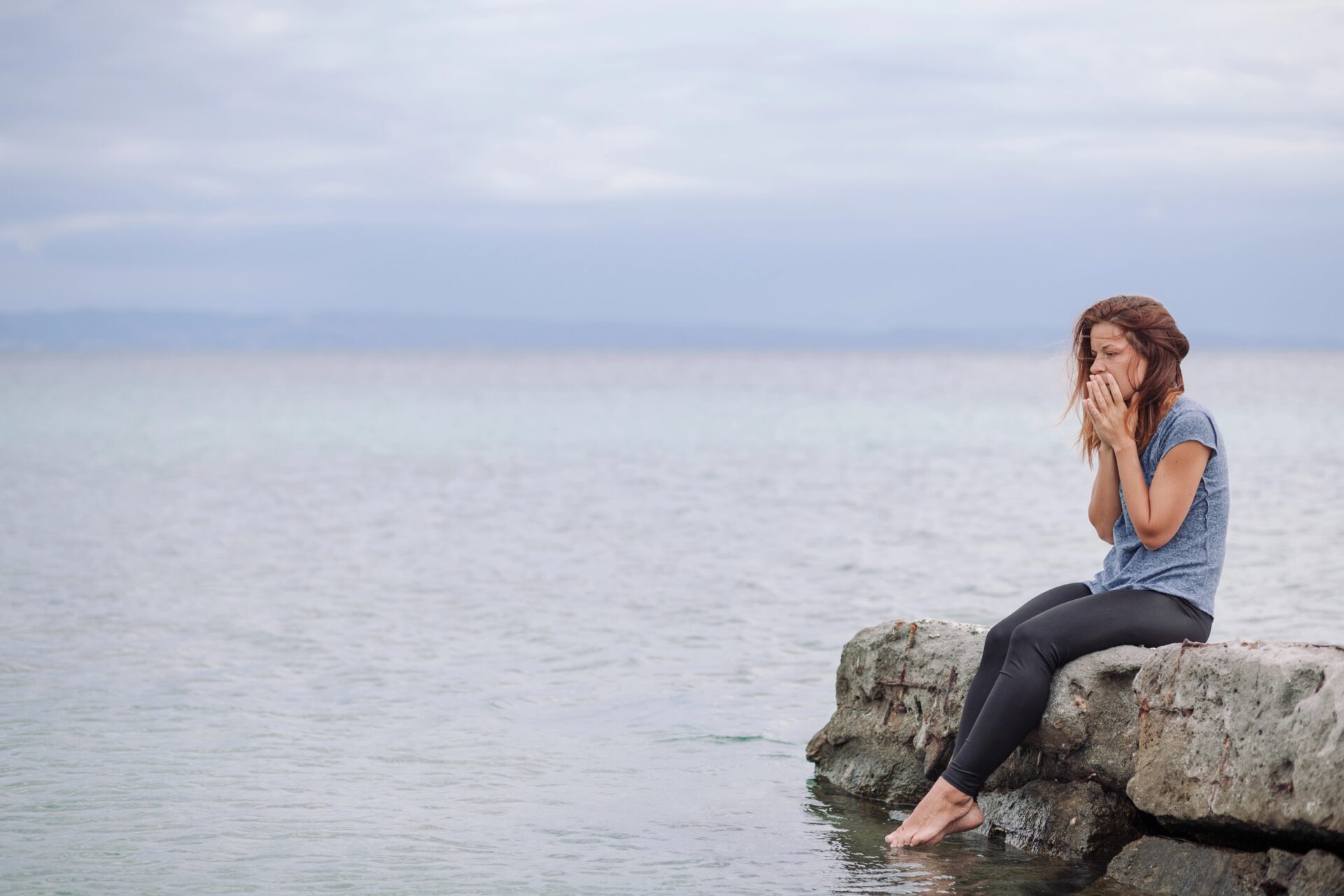 Woman sitting on rocks by water, experiencing emotional distress and seeking recovery.