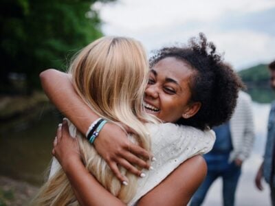 Two women embracing outdoors near water, smiling and happy.